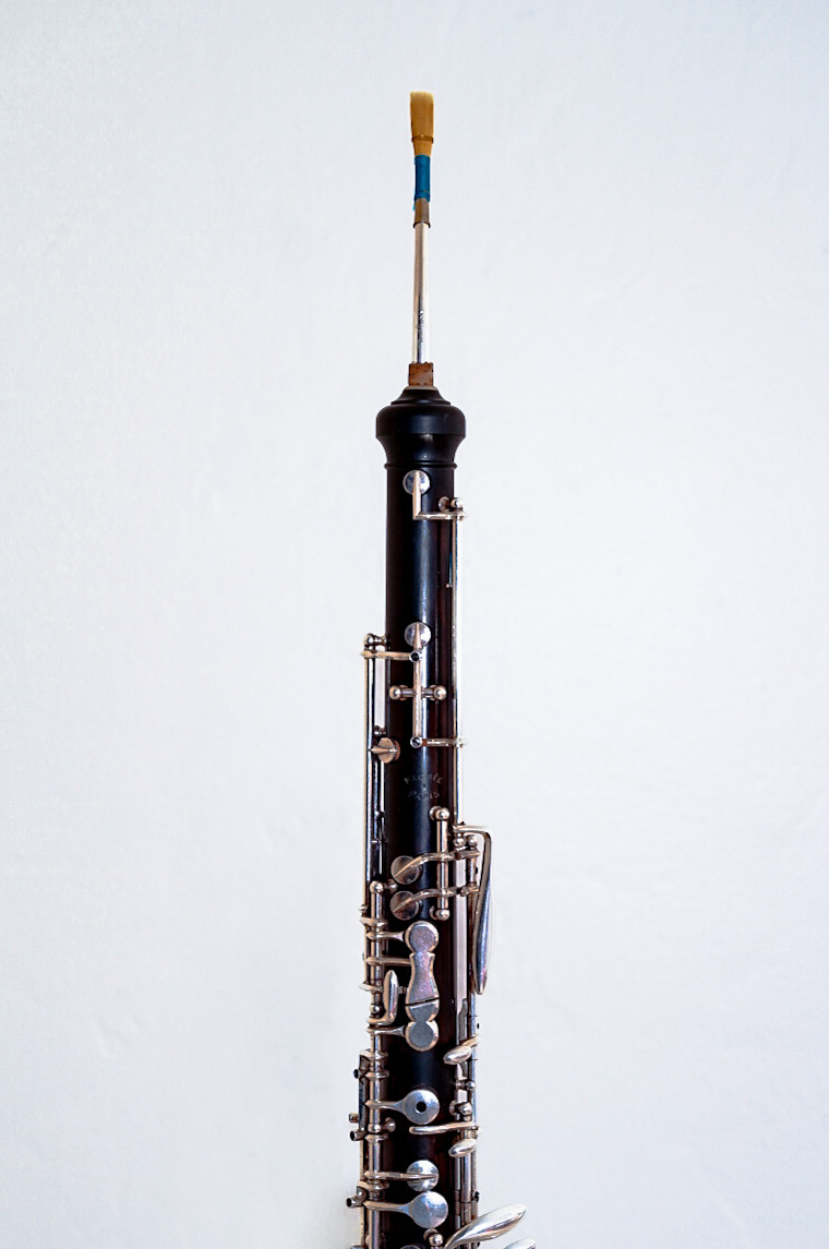 An english horn stands upright against a white wall. This instrument is known for its melancholy sound.